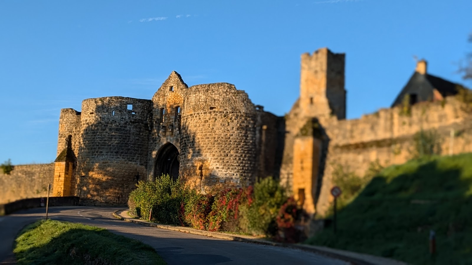 Porte fortifiée et tour de pierre dorées par le soleil couchant, route bordée d'arbustes