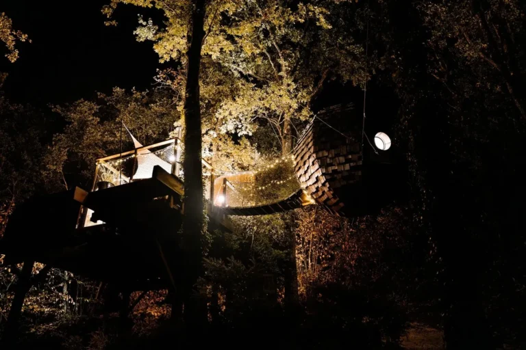 Le Lov'nid illuminé la nuit, cabane perchée et passerelle dans la forêt en Dordogne